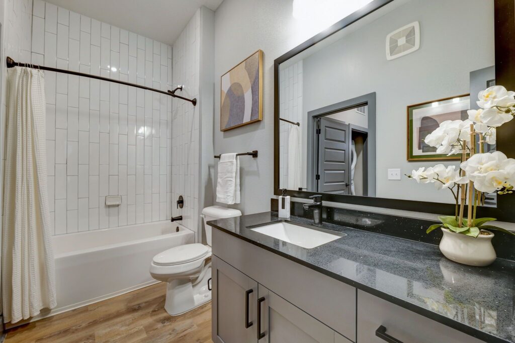 Modern bathroom with a white bathtub and shower curtain, black countertop sink, large mirror, toilet, towel rack, framed artworks, and a decorative orchid on the counter.