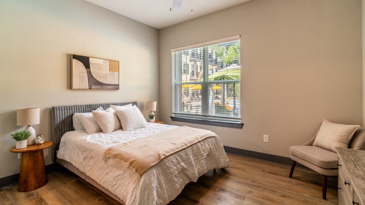 Bedroom with a queen bed, light brown bedding, wooden side table, painting above the headboard, large window with blinds, and hardwood flooring.