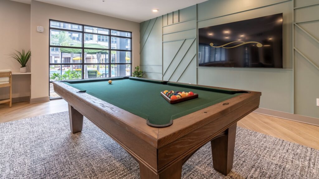 A pool table with a rack of balls is centered in a room with a large flat-screen TV on the wall and a window showing outdoor scenery.