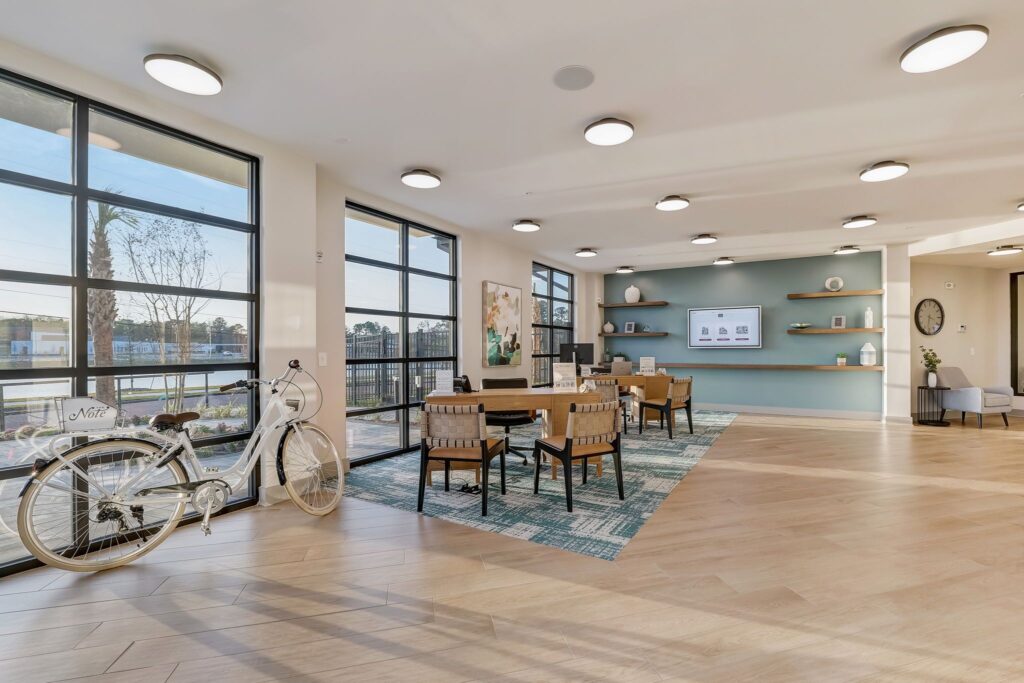 Bright open room with a bicycle, table, and chairs on a patterned rug. Shelves with decor on the right wall, large windows on the left. Ceiling lights illuminate the space.