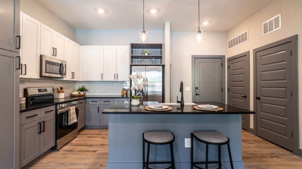 Modern kitchen with two-tone cabinets, stainless steel appliances, and a black countertop island with two stools. Hanging lights illuminate the space. Wood flooring throughout.