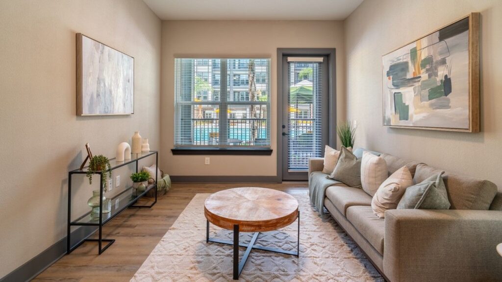 Modern living room with a gray sofa, round wooden coffee table, abstract wall art, and a glass console. Large window and door offer a view of an outdoor pool area.