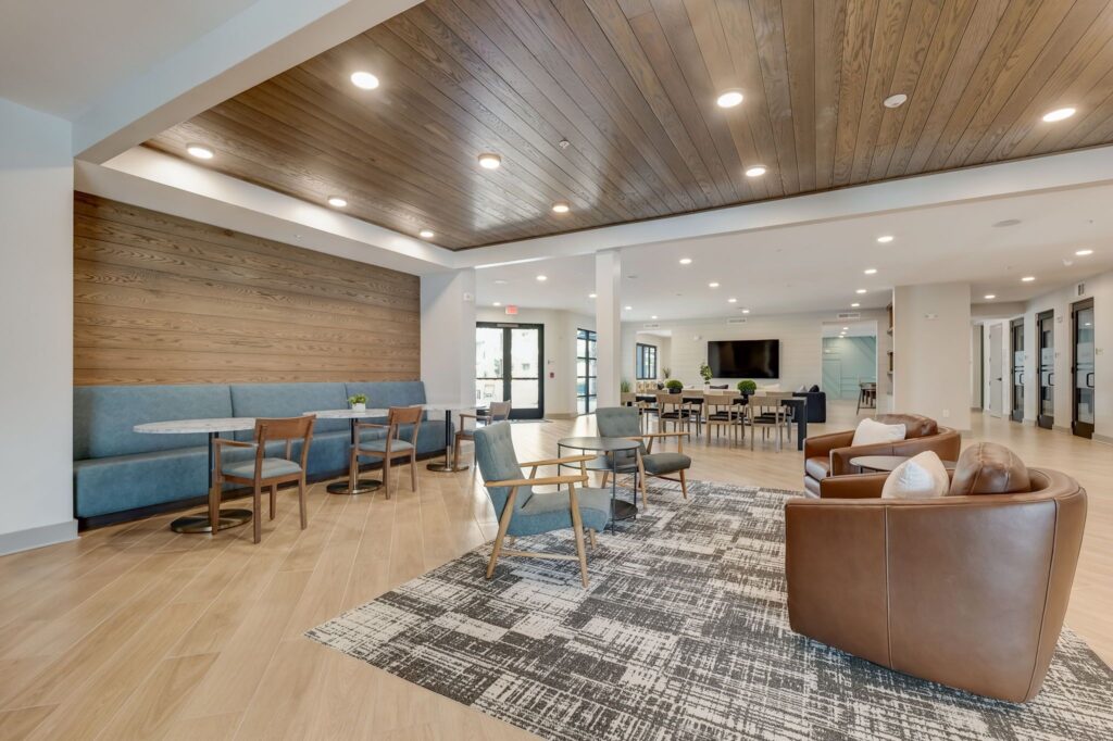 Modern communal lounge with wooden ceiling, seating areas including blue booths, brown armchairs, and a patterned rug. Walls feature wood paneling and large windows.