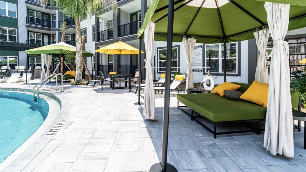 Poolside area with green lounge chairs, yellow and green umbrellas, and cabanas. An apartment building is in the background.