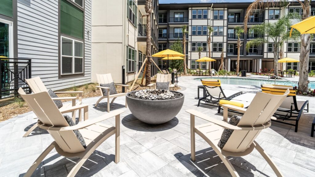 Outdoor patio with white chairs around a circular fire pit. Yellow umbrellas shade nearby tables. Modern apartment building stands in the background beside a small pool.