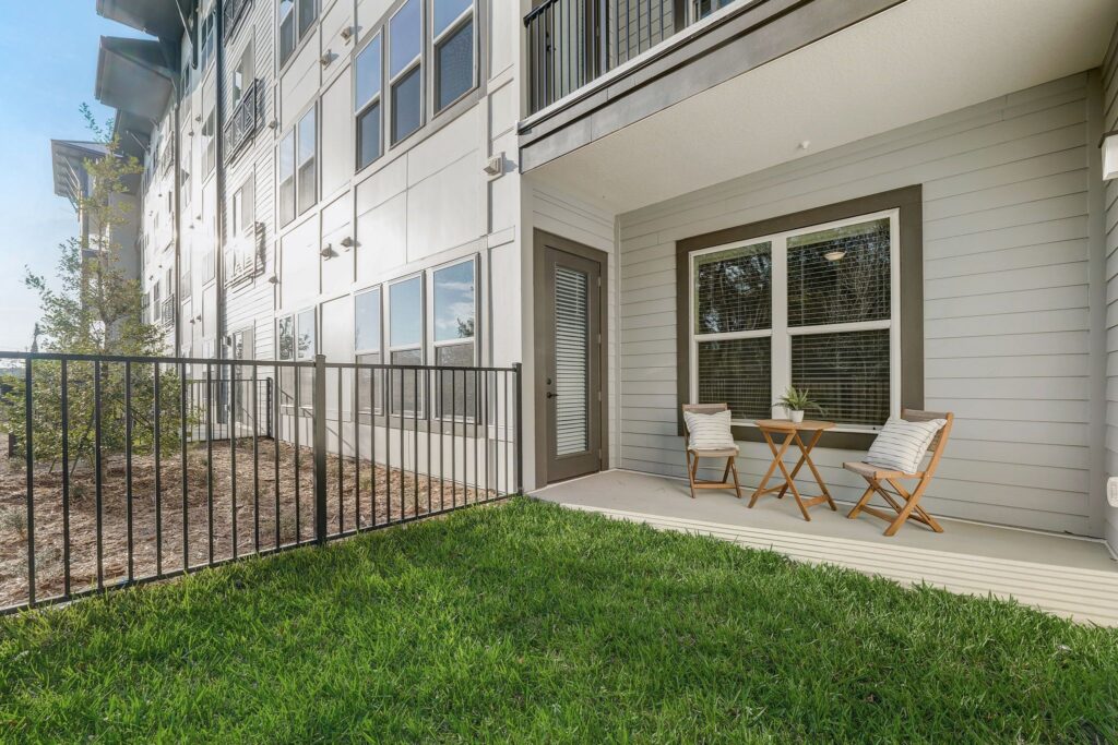 A ground-level patio with two wooden chairs and a small table near a modern building, enclosed by a black metal fence, and adjacent to a green lawn.