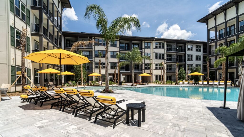 Apartment complex with a central swimming pool, surrounded by lounge chairs with yellow umbrellas, and palm trees under a clear blue sky.