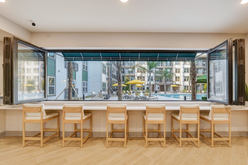 Counter seating with six wooden chairs overlooks a pool area through a large open window, with apartment buildings and outdoor furniture visible outside.