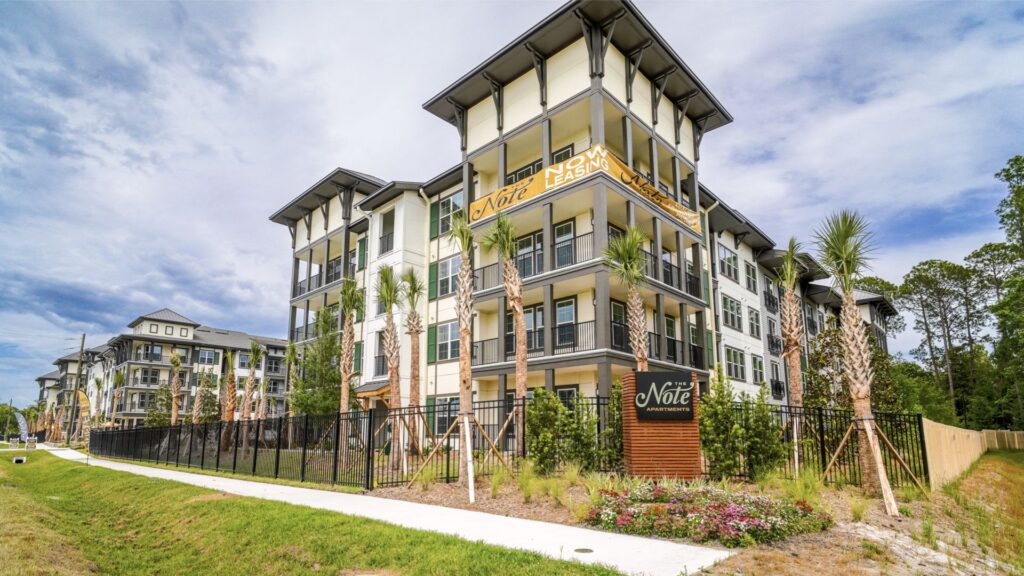 A four-story modern apartment building with balconies, surrounded by palm trees and a grassy area.