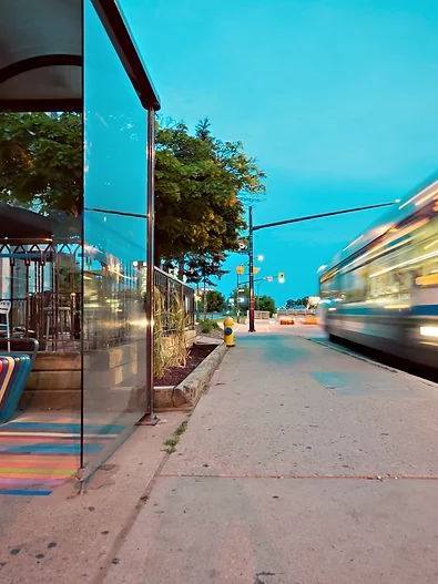 A city bus speeds past a glass shelter at a bus stop on an evening street, with trees and traffic lights in the background.