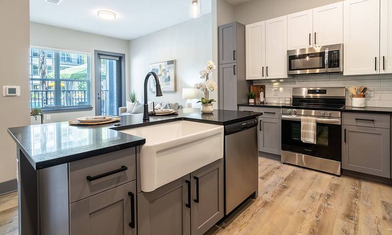 Modern kitchen with a farmhouse sink, stainless steel appliances, and gray cabinetry. Open layout with a view of the living area and large windows.