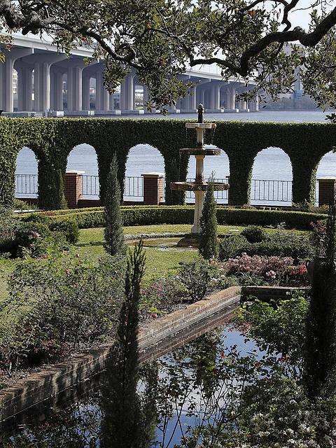 Garden with a fountain and pond, bordered by greenery. Arched hedge openings reveal a view of a river and a large bridge in the background.