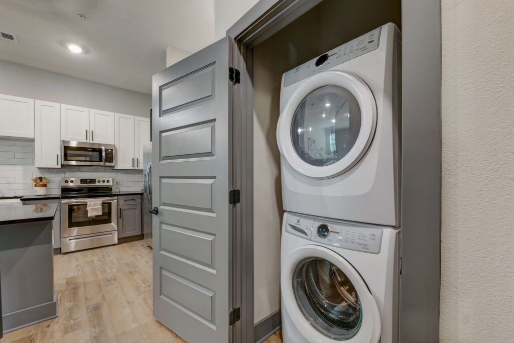 A compact laundry area with a white stacked washer and dryer inside a closet next to a kitchen featuring stainless steel appliances and light wood flooring.